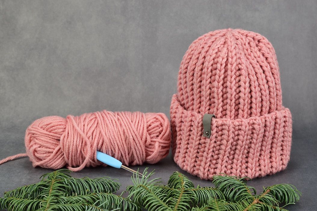 Close-up of a pink hand-knitted winter hat with matching yarn and crochet hook, styled with winter greenery on a dark background.