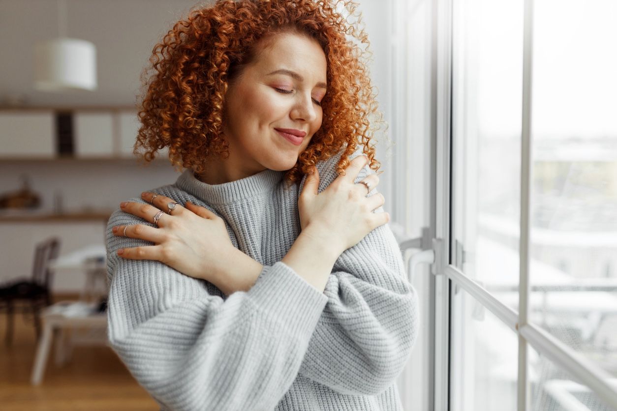 Smiling woman with curly red hair wearing a cozy gray sweater, hugging herself while standing by a bright window.
