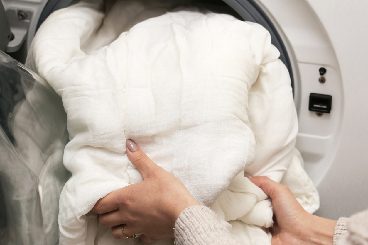 Person loading a white comforter into a washing machine before washing bulky bedding at home or the laundromat
